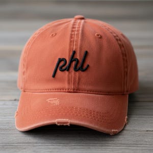 May include: A distressed orange baseball cap with the embroidered letters "phl" in black script. The cap has a curved brim and a slightly faded appearance, with distressed details around the brim.