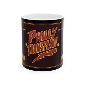 May include: A dark brown ceramic mug featuring the text PHILLY TRANSPLANT in a distressed yellow and red font. A yellow lightning bolt graphic is positioned below the text. The words CAN'T WE ALL GET ALONG? are printed in small yellow letters.