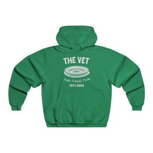 Op de afbeelding: Een groene hoodie met de tekst "THE VET" boven een stadionafbeelding. Daaronder staat "Pride, Pretzels, Prison 1971-2003". De sweatshirt heeft een voorzak en een capuchon met trekkoord.