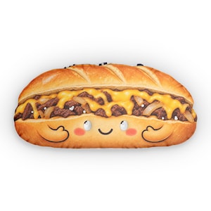 Può includere: Un cuscino in peluche a forma di panino cheesesteak con pane dorato, carne marrone scuro, formaggio giallo fuso e cipolle saltate. La parte inferiore del pane presenta un volto da cartone animato stampato con grandi occhi e guance rosa.