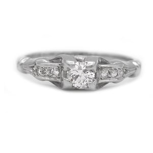 May include: A silver-toned metal ring featuring a central round-cut clear gemstone held in a four-prong setting. The band is accented with three smaller clear stones on each side, embedded within the metal shank, creating a continuous design.