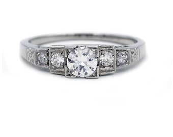 May include: A silver ring with a large, round, clear diamond center stone, flanked by smaller diamonds. The band features intricate engraved detailing. The ring is set against a white background.