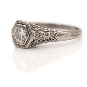 Puede incluir: Un anillo de compromiso de filigrana de plata con un solo diamante redondo en un engaste hexagonal.