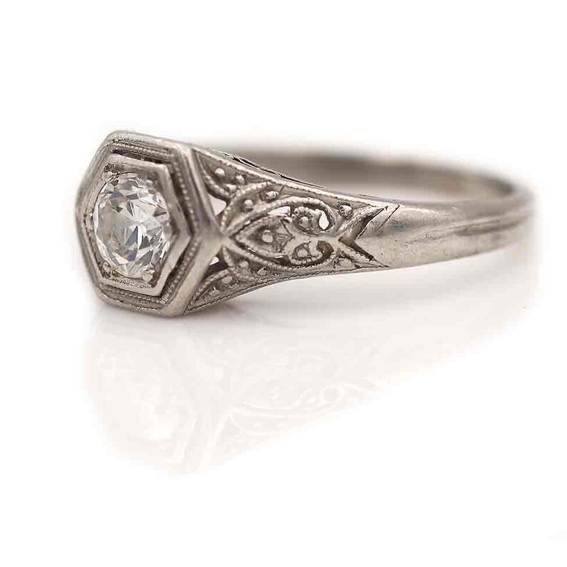 Puede incluir: Un anillo de compromiso de filigrana de plata con un solo diamante redondo en un engaste hexagonal.