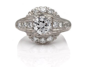 May include: A platinum engagement ring with a round brilliant cut diamond center stone surrounded by smaller diamonds. The ring has a delicate, vintage-inspired design.