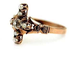Puede incluir: Un anillo de oro con un dise&ntilde;o de cruz engastado con una piedra grande transparente y perlas blancas m&aacute;s peque&ntilde;as.