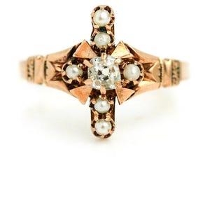 Puede incluir: Un anillo de oro con un diamante central y detalles de perlas. El anillo est&aacute; engastado en forma de cruz con un dise&ntilde;o delicado.