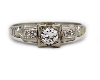 May include: A vintage-style engagement ring with a large, round, clear diamond as the centerpiece. The ring has a silver-colored band with smaller accent diamonds on either side of the main stone. The setting has a square shape around the main diamond.