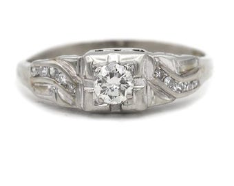 May include: A silver-toned ring with a central, round-cut diamond. The ring features a square setting for the main stone and smaller diamonds on each side. The band has a decorative, Art Deco-inspired design.