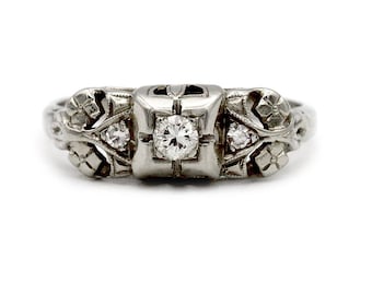 May include: A vintage-style ring with a central, round-cut diamond, flanked by two smaller diamonds. The ring is crafted from a light-colored metal, possibly platinum or white gold, and features intricate floral and geometric designs. The ring's band tapers towards the back.