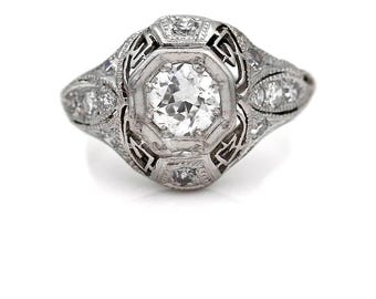 May include: A vintage platinum engagement ring with a large, round, brilliant-cut diamond surrounded by smaller diamonds in a geometric design.
