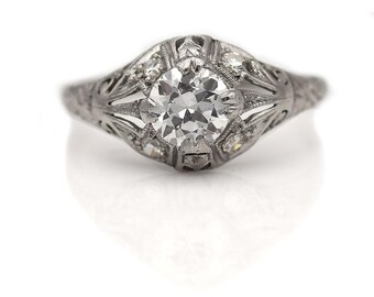 May include: A silver ring with a round, clear diamond center stone and smaller diamonds surrounding it. The ring has a delicate, intricate design.