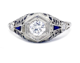 May include: A vintage engagement ring with a round brilliant cut diamond set in a hexagonal bezel. The ring features a filigree design with blue enamel accents.
