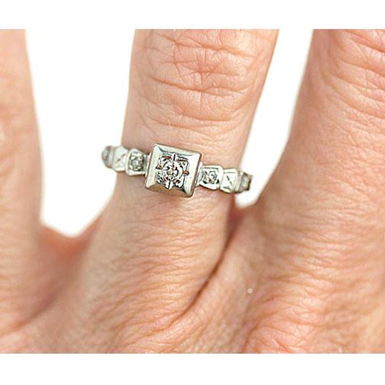 May include: A silver ring with a square-shaped center stone and smaller stones set around the band.
