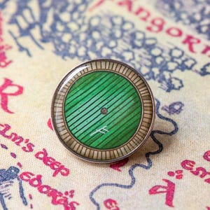 Op de afbeelding: Een ronde speld met een groen deurontwerp, horizontale houten planken en een rond raam in het midden. De speld heeft een metalen rand met een radiaal patroon en een klein wit symbool gegraveerd op het onderste deel van het groene vlak.