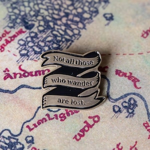 Puede incluir: Un pin de metal plateado con una cinta de listón negro que dice "Not all those who wander are lost." El pin está en un mapa de la Tierra Media.