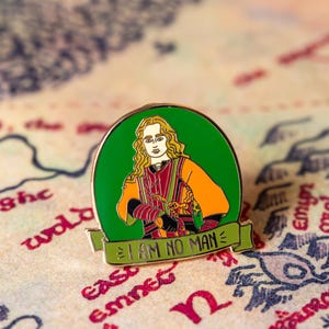 Könnte beinhalten: Eine grüne und goldene Emaille-Anstecknadel mit einem Cartoon-Bild einer Person mit langen blonden Haaren und einer Krone. Die Person trägt eine rote und orangefarbene Robe und hält ein Schwert. Der Text "I AM NO MAN" ist auf einem grünen Banner unter der Person geschrieben.