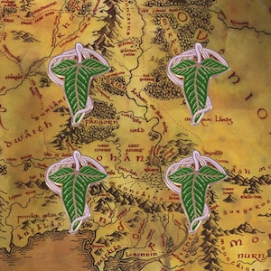 Puede incluir: Cuatro pines verdes con forma de hoja con detalles blancos y dorados sobre un fondo de mapa. Los pines tienen un diseño detallado de hoja y un borde decorativo. El mapa muestra nombres de lugares y detalles geográficos.