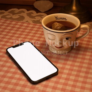 May include: A black smartphone with a blank white screen rests on a red and white checkered tablecloth. Beside it is a whimsical teacup filled with tea, featuring a winking face design and the word "Yumm." The scene is lit with warm, inviting light.