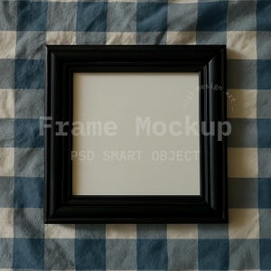 May include: A square black picture frame with a white interior, set against a blue and white checkered fabric background. The frame has a thick, beveled edge. Text on the frame reads "Frame Mockup" and "PSD SMART OBJECT".