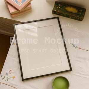 May include: A black framed mockup with a white mat, a stack of pink tarot books, and a green and gold decorative box. A small green cup sits on a white embroidered cloth. The words "Frame Mockup" and "PSD SMART OBJECT" are visible.