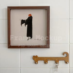 May include: A framed vintage illustration of a person in a long black coat and orange hat. The artwork is in a dark brown wooden frame. Below the frame is a wooden key holder with three hooks and a key.