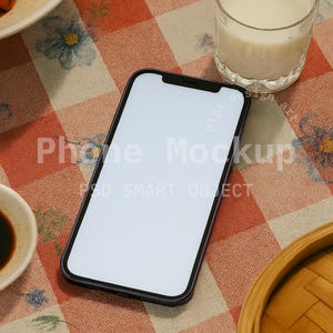 May include: A smartphone with a blank white screen rests on a red and white checkered tablecloth. A glass of milk, a small bowl of sauce, and a wooden steamer basket are also on the table. The words "Phone Mockup" and "PSD SMART OBJECT" are visible.