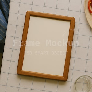Könnte beinhalten: Ein rechteckiger Holzbilderrahmen mit hellem Innenraum. Der Rahmen hat abgerundete Ecken und liegt auf einer karierten Tischdecke. Der Text "Frame Mockup" und "PSD SMART OBJECT" sind auf dem Rahmen zu sehen.