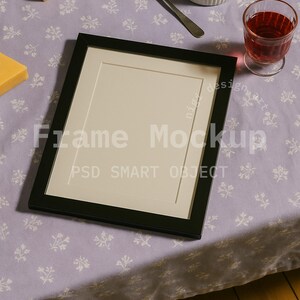 May include: A black picture frame mockup with the text "Frame Mockup PSD SMART OBJECT" is displayed on a lavender floral tablecloth. Cheese, a spoon with cheese, a small bowl of cheese, and a glass of red wine are also present.