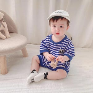 Peut inclure: Un bébé portant un ensemble rayé bleu et blanc et une casquette de baseball blanche. L'ensemble a un petit motif sur la poitrine. Le bébé porte également des chaussettes blanches avec des détails noirs. Le bébé tient un jouet en bois.