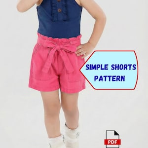 May include: A child wearing a navy blue top with a ruffled neckline and pink shorts with a bow. The shorts have pockets and a high waist. Text on the image reads "SIMPLE SHORTS PATTERN". A PDF icon is in the lower right corner.