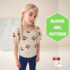 Puede incluir: Una blusa beige de manga corta con un estampado repetitivo de cerezas rojas y hojas verdes. La blusa tiene un escote cuadrado y mangas abullonadas. Un gráfico hexagonal verde muestra el texto BLOUSE GIRL PATTERN.