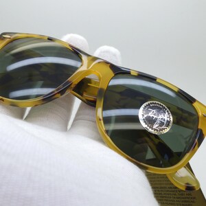 May include: Tortoiseshell sunglasses with dark lenses. The frames are a mix of yellow and brown, with a classic shape. A silver sticker is on the lens. The sunglasses are held by a gloved hand.