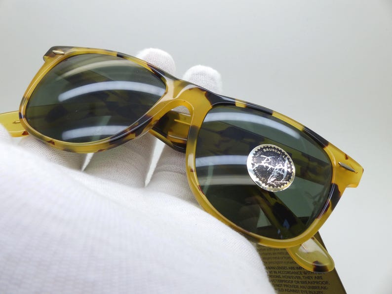May include: Tortoiseshell sunglasses with dark lenses. The frames are a mix of yellow and brown, with a classic shape. A silver sticker is on the lens. The sunglasses are held by a gloved hand.