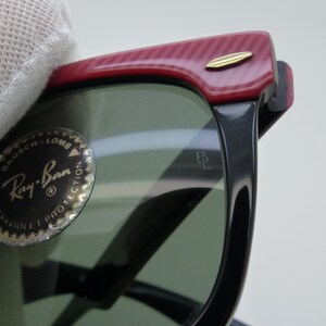 Puede incluir: Primer plano de gafas de sol Ray-Ban con montura negra y lentes verdes. La parte superior de la montura es roja con un patr&oacute;n texturizado y un acento dorado. El logotipo de Ray-Ban es visible en la lente.