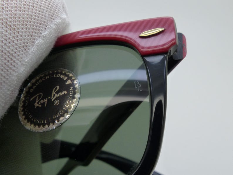 Puede incluir: Primer plano de gafas de sol Ray-Ban con montura negra y lentes verdes. La parte superior de la montura es roja con un patr&oacute;n texturizado y un acento dorado. El logotipo de Ray-Ban es visible en la lente.