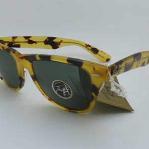 May include: Tortoiseshell sunglasses with dark green lenses. The frames have a classic design with a speckled pattern in shades of yellow and brown. A tag is attached to the arm.