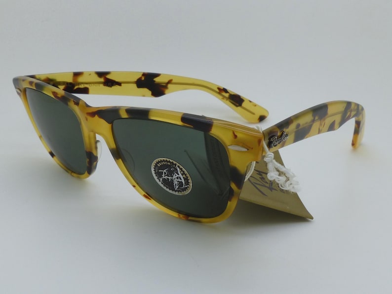 May include: Tortoiseshell sunglasses with dark green lenses. The frames have a classic design with a speckled pattern in shades of yellow and brown. A tag is attached to the arm.