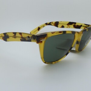 May include: Tortoiseshell-patterned sunglasses with dark green lenses. The frames are a mix of yellow and brown, with the Ray-Ban logo on the temple. These sunglasses offer a classic, stylish look.