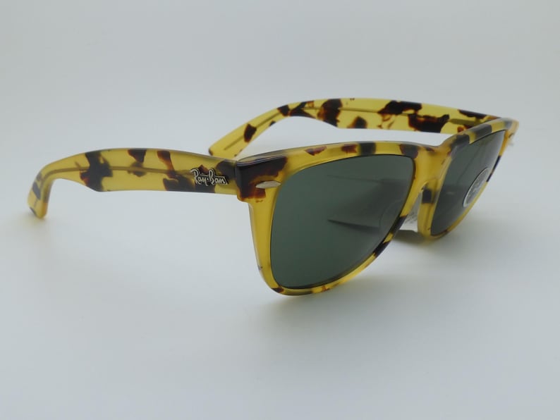 May include: Tortoiseshell-patterned sunglasses with dark green lenses. The frames are a mix of yellow and brown, with the Ray-Ban logo on the temple. These sunglasses offer a classic, stylish look.