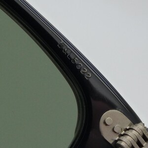 Puede incluir: Primer plano de una montura de gafas de sol negra con lente verde. El texto "B&L 5022" est&aacute; grabado en la varilla. La bisagra y la varilla son plateadas. Las gafas de sol est&aacute;n en &aacute;ngulo, mostrando los detalles de la montura.