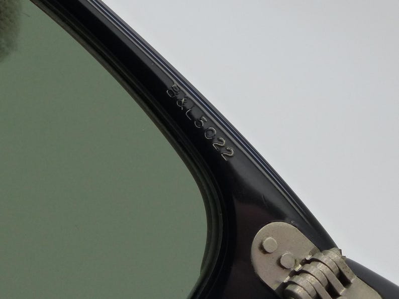 Puede incluir: Primer plano de una montura de gafas de sol negra con lente verde. El texto "B&L 5022" est&aacute; grabado en la varilla. La bisagra y la varilla son plateadas. Las gafas de sol est&aacute;n en &aacute;ngulo, mostrando los detalles de la montura.