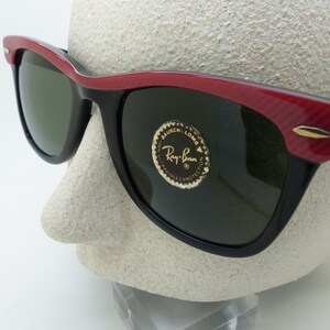 Puede incluir: Gafas de sol negras con montura roja y negra. Las gafas tienen lentes verde oscuro y un acento dorado en las patillas. El logotipo de Ray-Ban es visible en la lente.