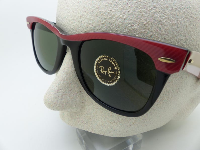 Puede incluir: Gafas de sol negras con montura roja y negra. Las gafas tienen lentes verde oscuro y un acento dorado en las patillas. El logotipo de Ray-Ban es visible en la lente.