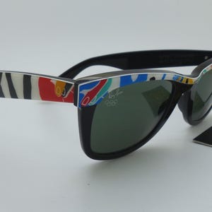 May include: Black Ray-Ban sunglasses with a colorful pattern on the arms. The sunglasses have dark lenses and a tag that reads "Ray-Ban Olympic Games". The arms feature a mix of red, white, blue, and yellow designs.
