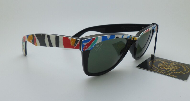 May include: Black Ray-Ban sunglasses with a colorful pattern on the arms. The sunglasses have dark lenses and a tag that reads "Ray-Ban Olympic Games". The arms feature a mix of red, white, blue, and yellow designs.