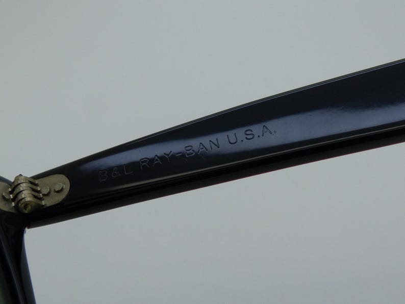Puede incluir: Primer plano de una montura de gafas negra con el texto "B&L RAY-BAN U.S.A." impreso en la patilla. La bisagra es visible, con metal plateado. La montura es brillante y refleja la luz.