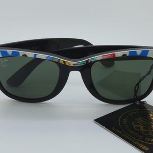 May include: Black Ray-Ban sunglasses with green lenses and a colorful patterned design on the frame. The sunglasses have the Ray-Ban logo and a tag that reads "Official Licensed Sunglasses".