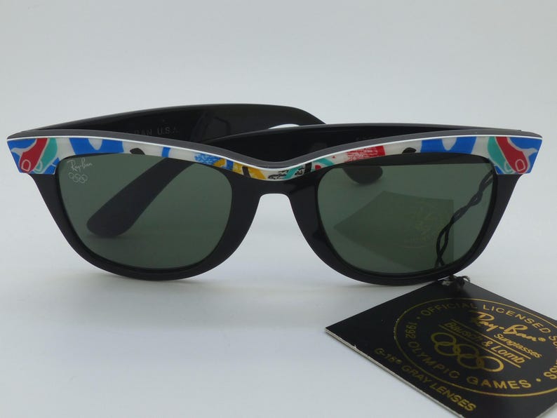 May include: Black Ray-Ban sunglasses with green lenses and a colorful patterned design on the frame. The sunglasses have the Ray-Ban logo and a tag that reads "Official Licensed Sunglasses".