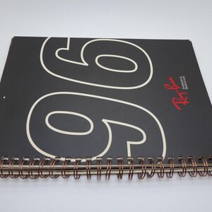 Puede incluir: Un cuaderno negro con espiral y el n&uacute;mero "96" en blanco en la portada. El logotipo de Ray-Ban es rojo. La encuadernaci&oacute;n es de color cobre.